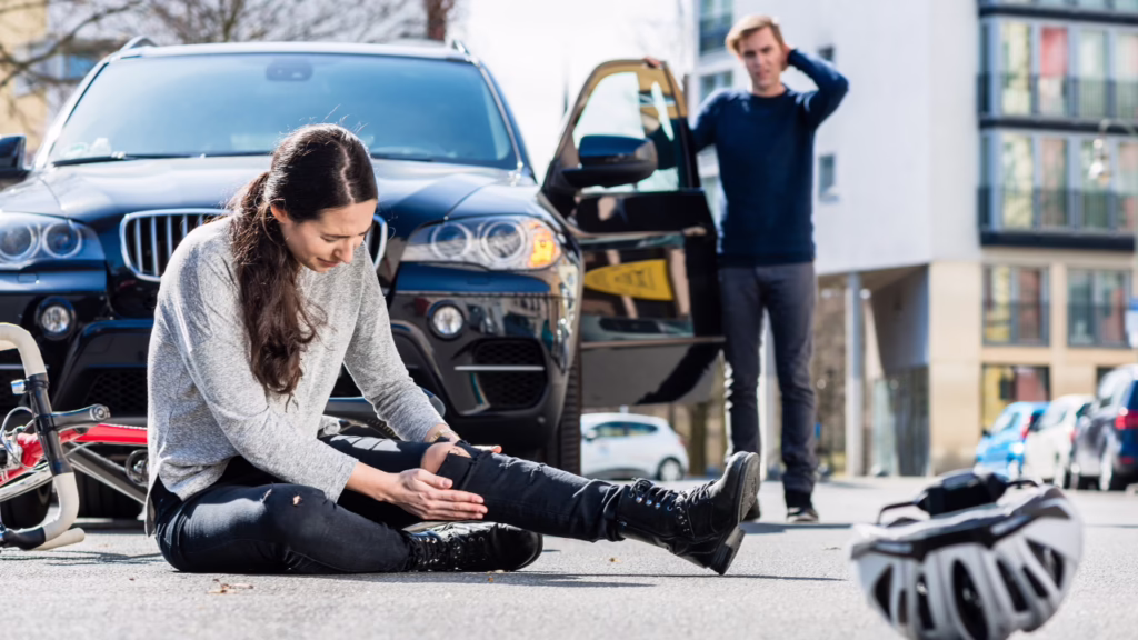 Nevada Bicycle Laws: What Cyclists Need to Know After a Crash 1 A woman sits injured on the street beside a bicycle, while a concerned man stands by an open car door in the background. © Marathon Law Group
